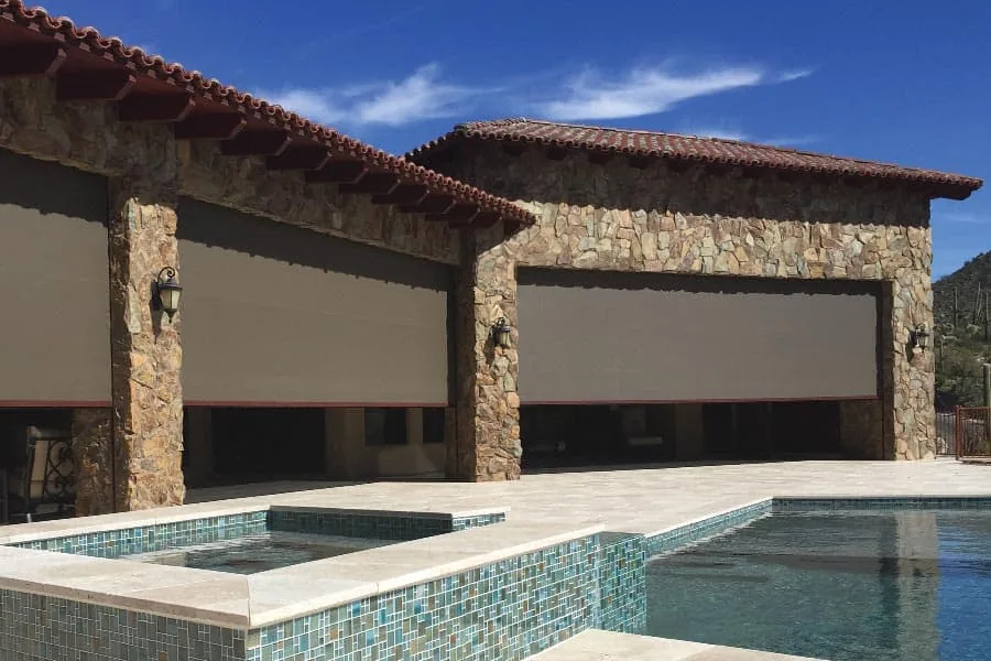 Poolside Stone Columns, two wide solar shades lowered between stone columns under a tile roof above a turquoise pool and spa
